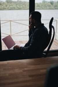 A person working remotely on a laptop by a large window with a lakeside view, conveying a sense of contemplation.