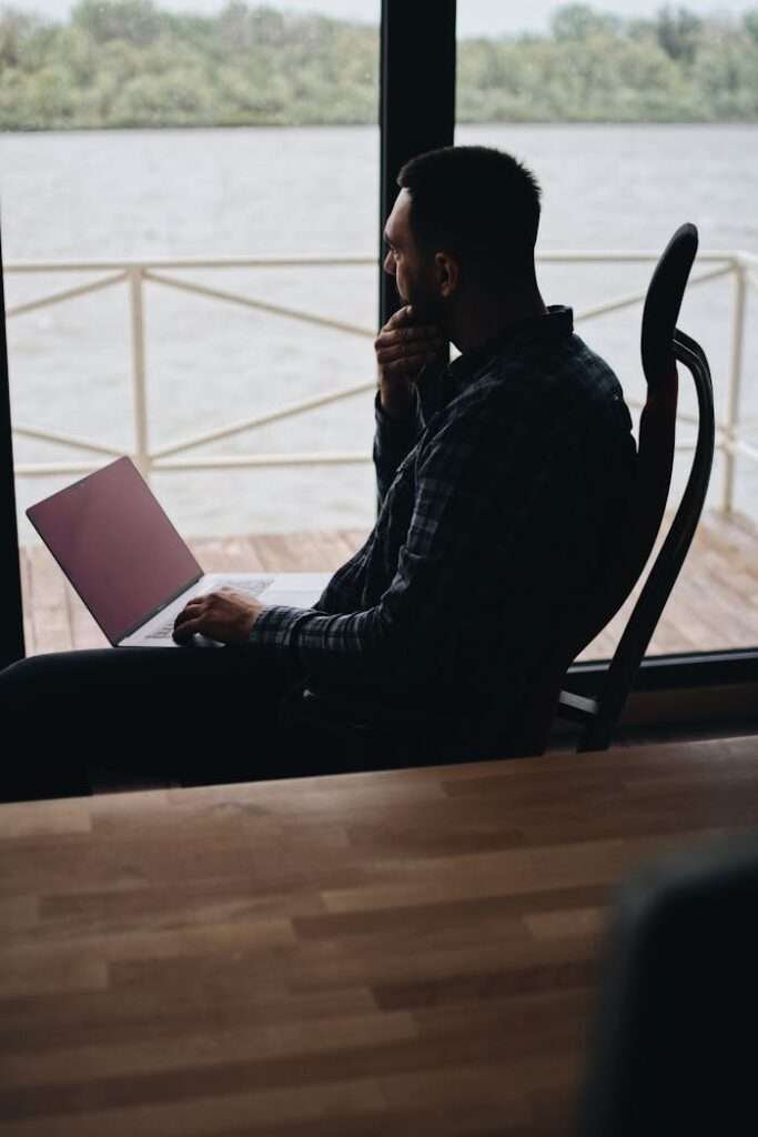 A person working remotely on a laptop by a large window with a lakeside view, conveying a sense of contemplation.